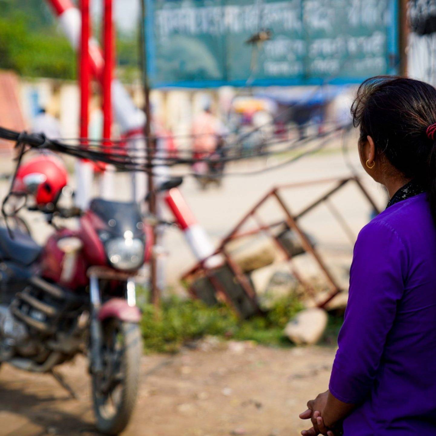 A woman facing away from the camera, stands in front of a motorcycle.