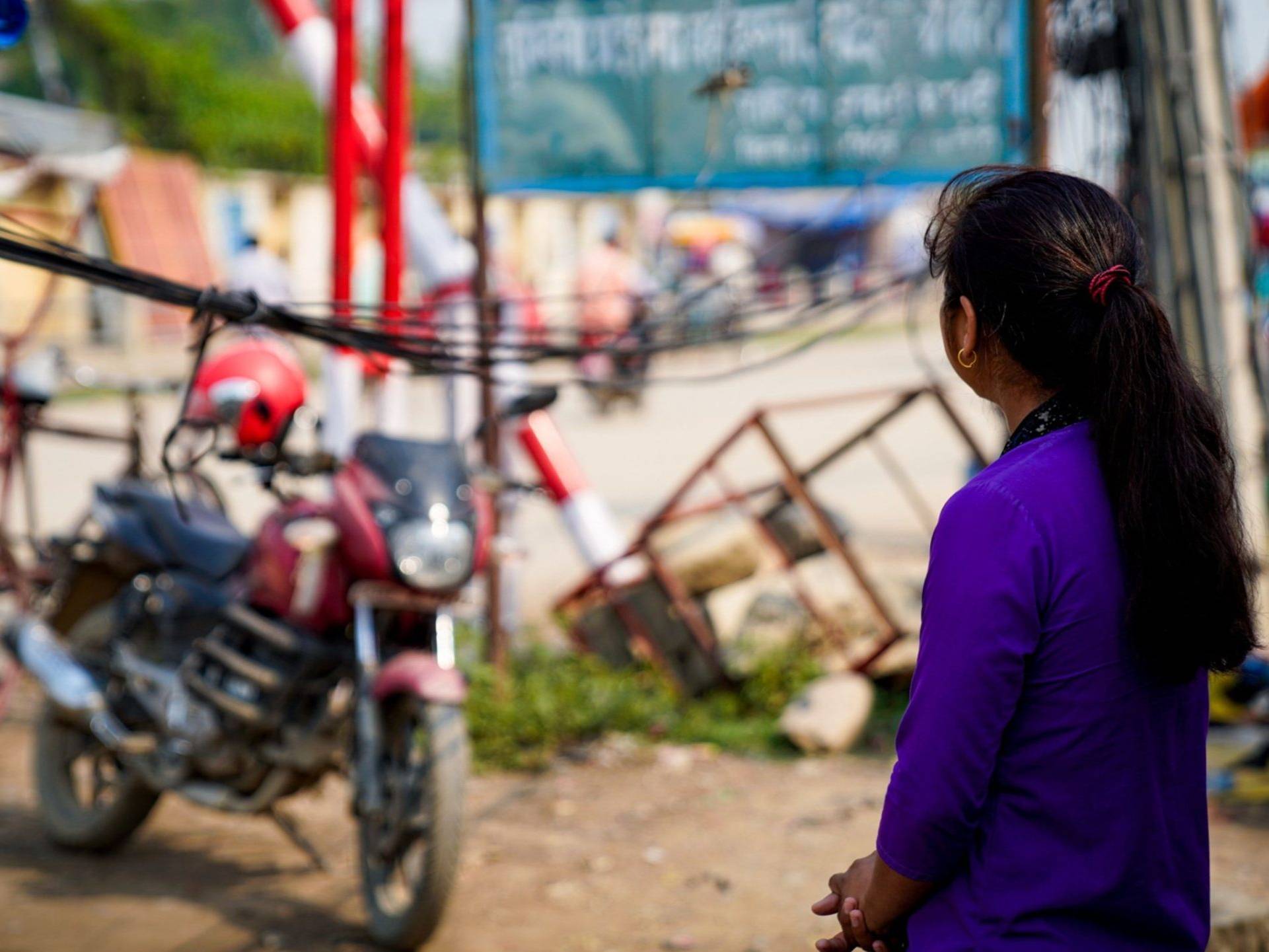 A woman facing away from the camera, stands in front of a motorcycle.