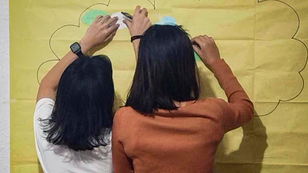 Two women are collaboratively posting on a wall, featuring a large cloud in the design.