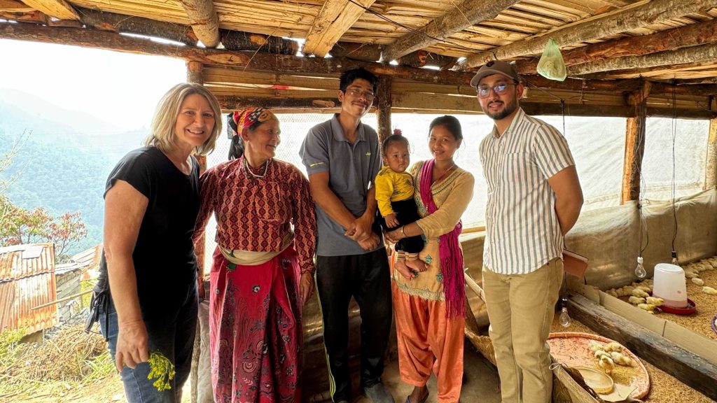 A diverse group of people stands together in front of a rustic hut, smiling and engaging with one another.