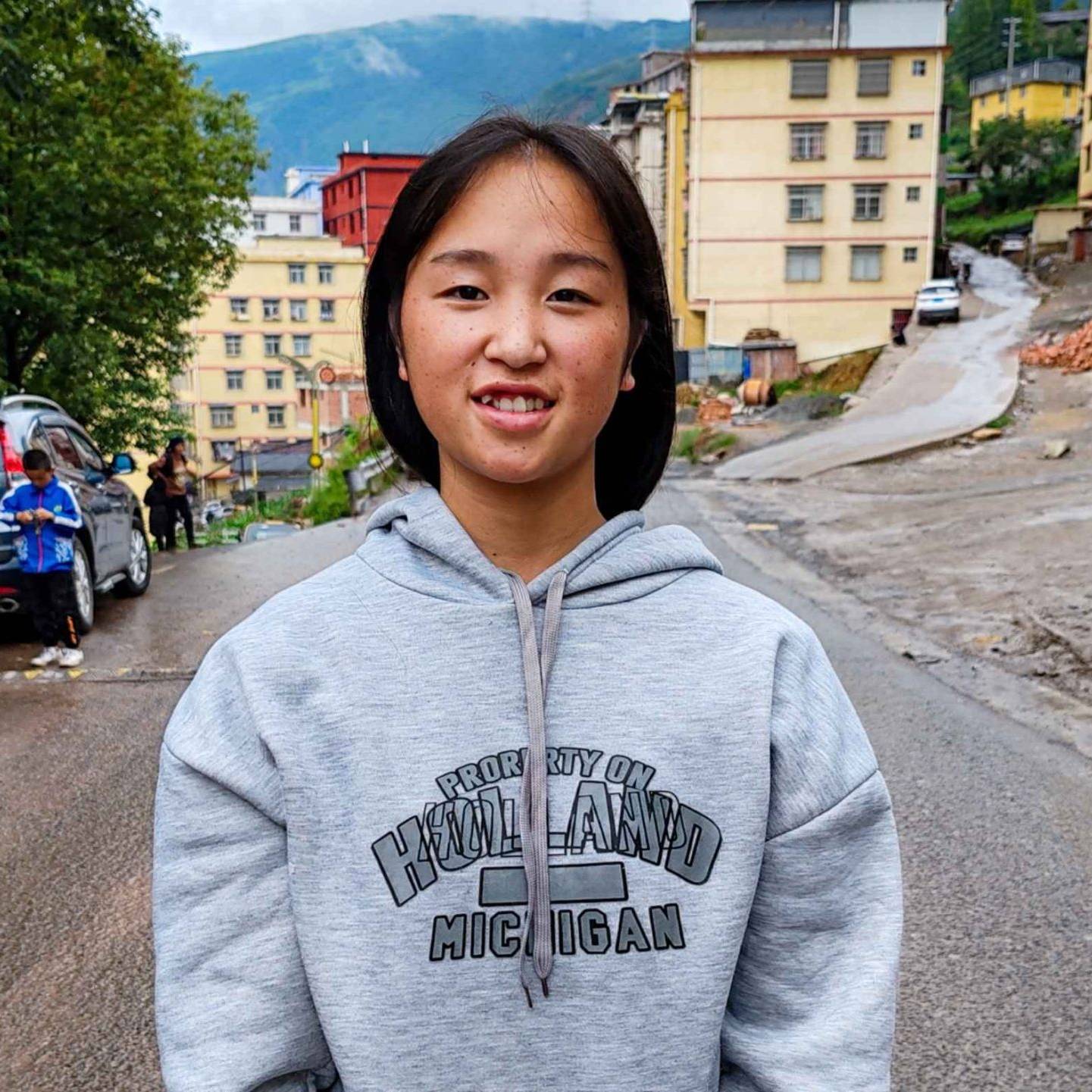 A young woman in a hoodie stands on a street, looking confidently at the camera with urban surroundings in the background.