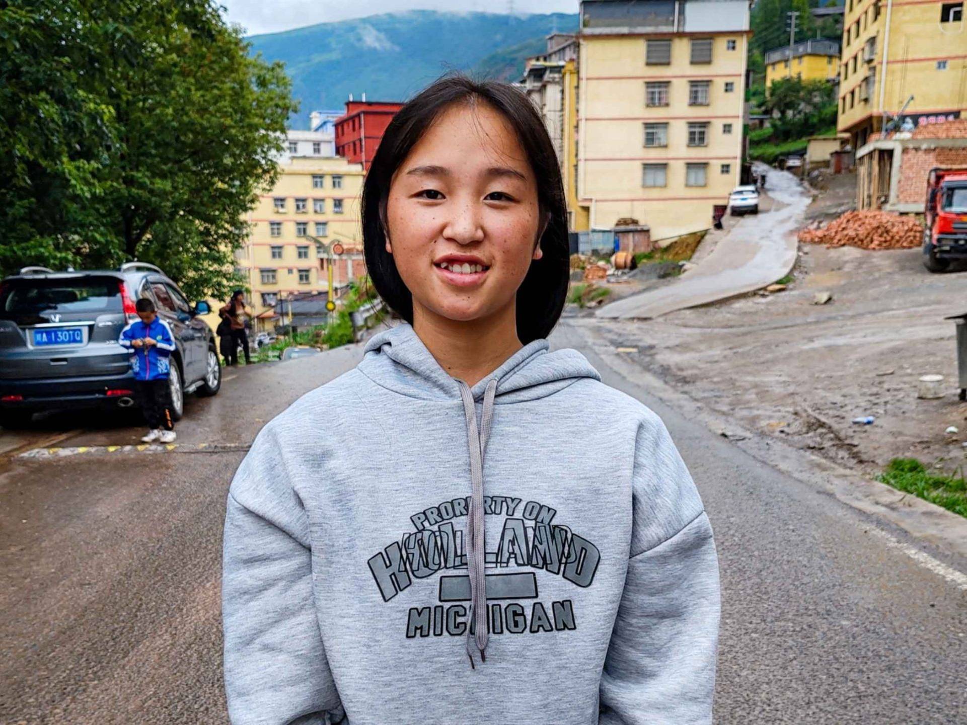 A young woman in a hoodie stands on a street, looking confidently at the camera with urban surroundings in the background.