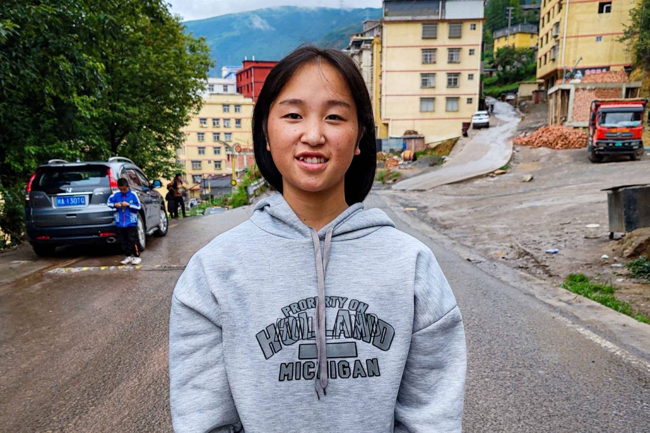 A young woman in a hoodie stands on a street, looking confidently at the camera with urban surroundings in the background.