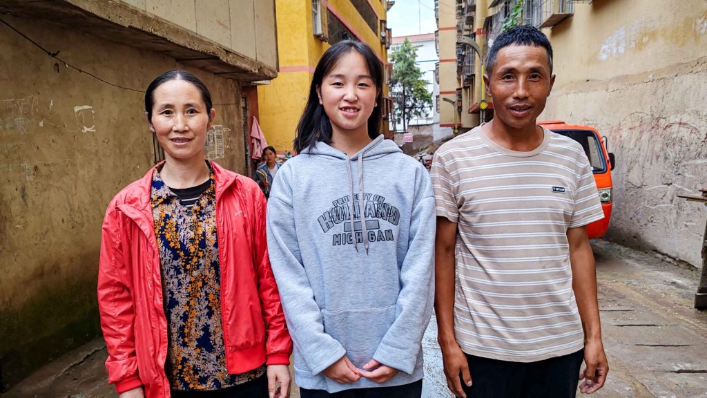 Three individuals standing closely together in a narrow alley, surrounded by brick walls and dim lighting.