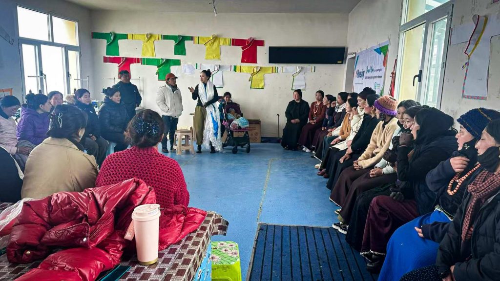 In a tailoring class, a woman and a man stand while a group of people sit around them, engaged in learning and discussion.