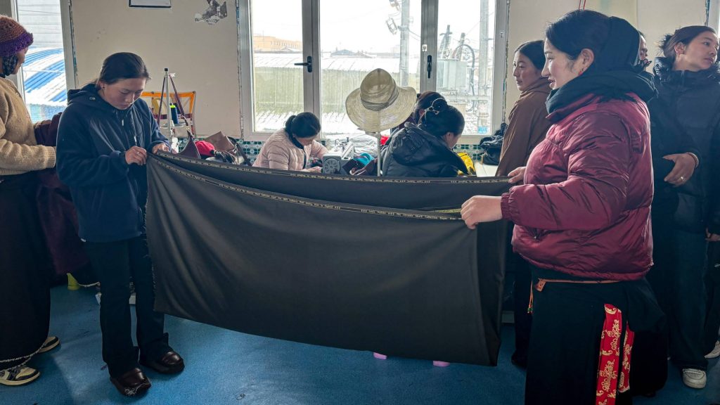 A group of people in a tailoring class with two women in the middle folding a large piece of cloth.