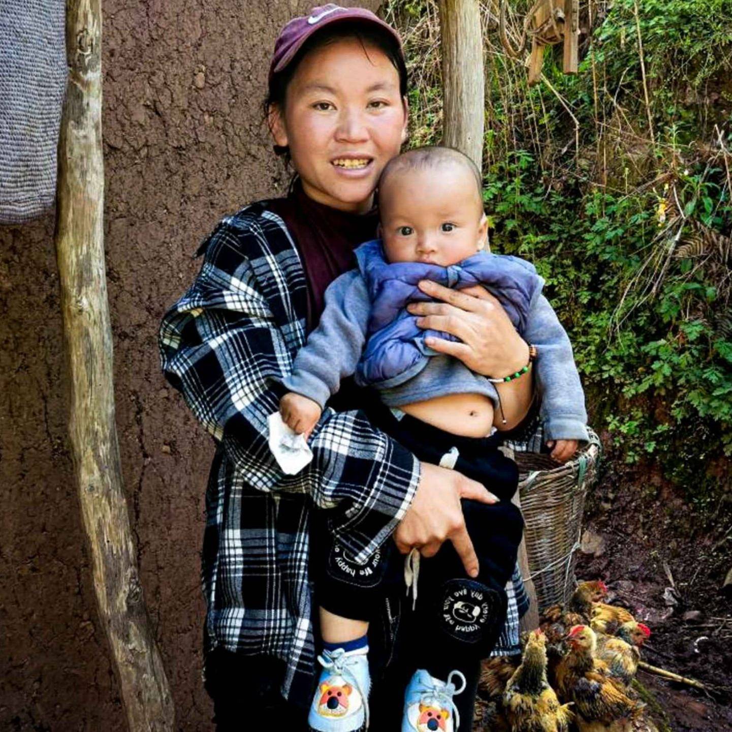 A woman cradles a baby in front of a rustic hut, showcasing a tender moment in a natural setting.
