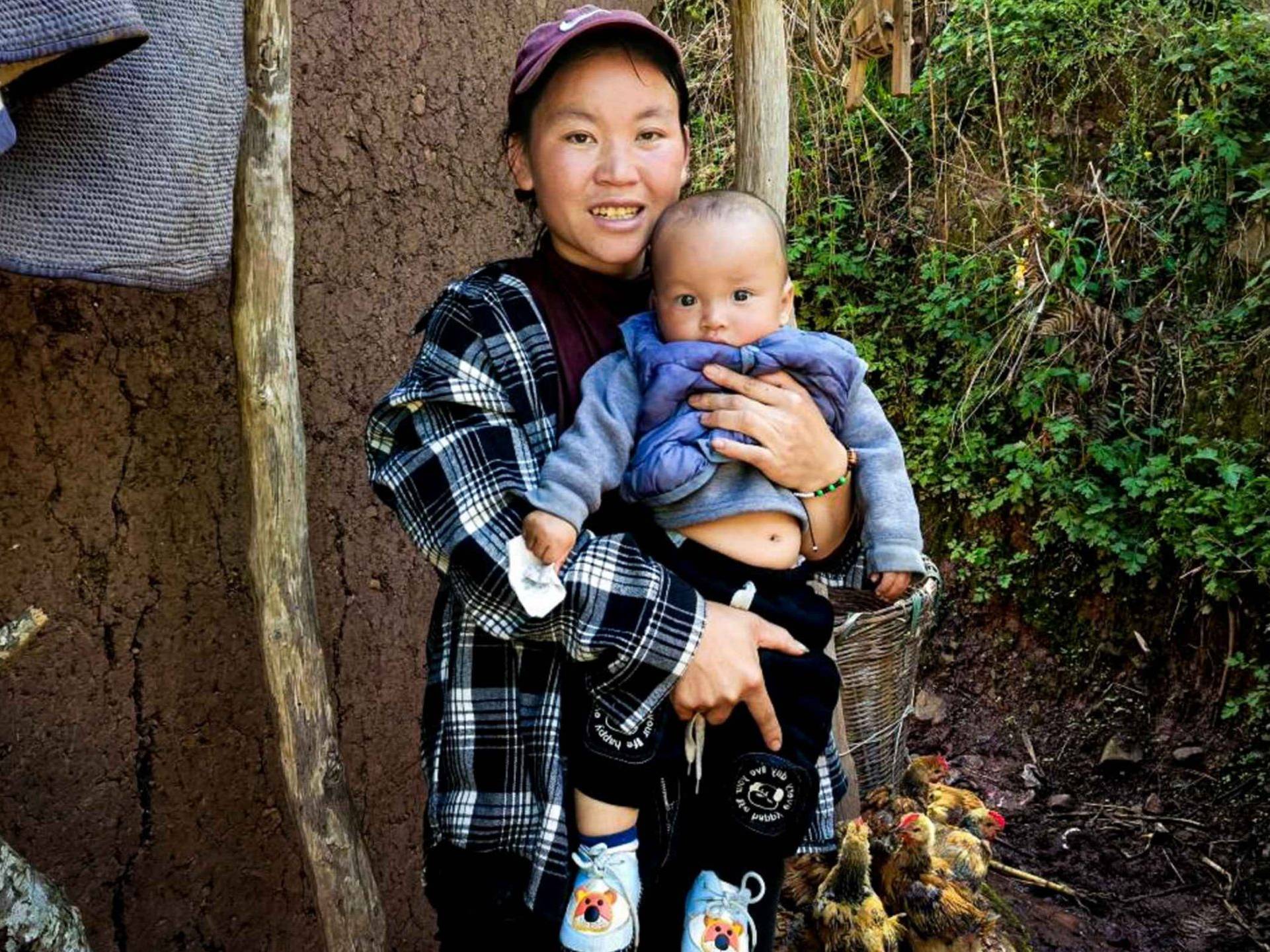 A woman cradles a baby in front of a rustic hut, showcasing a tender moment in a natural setting.