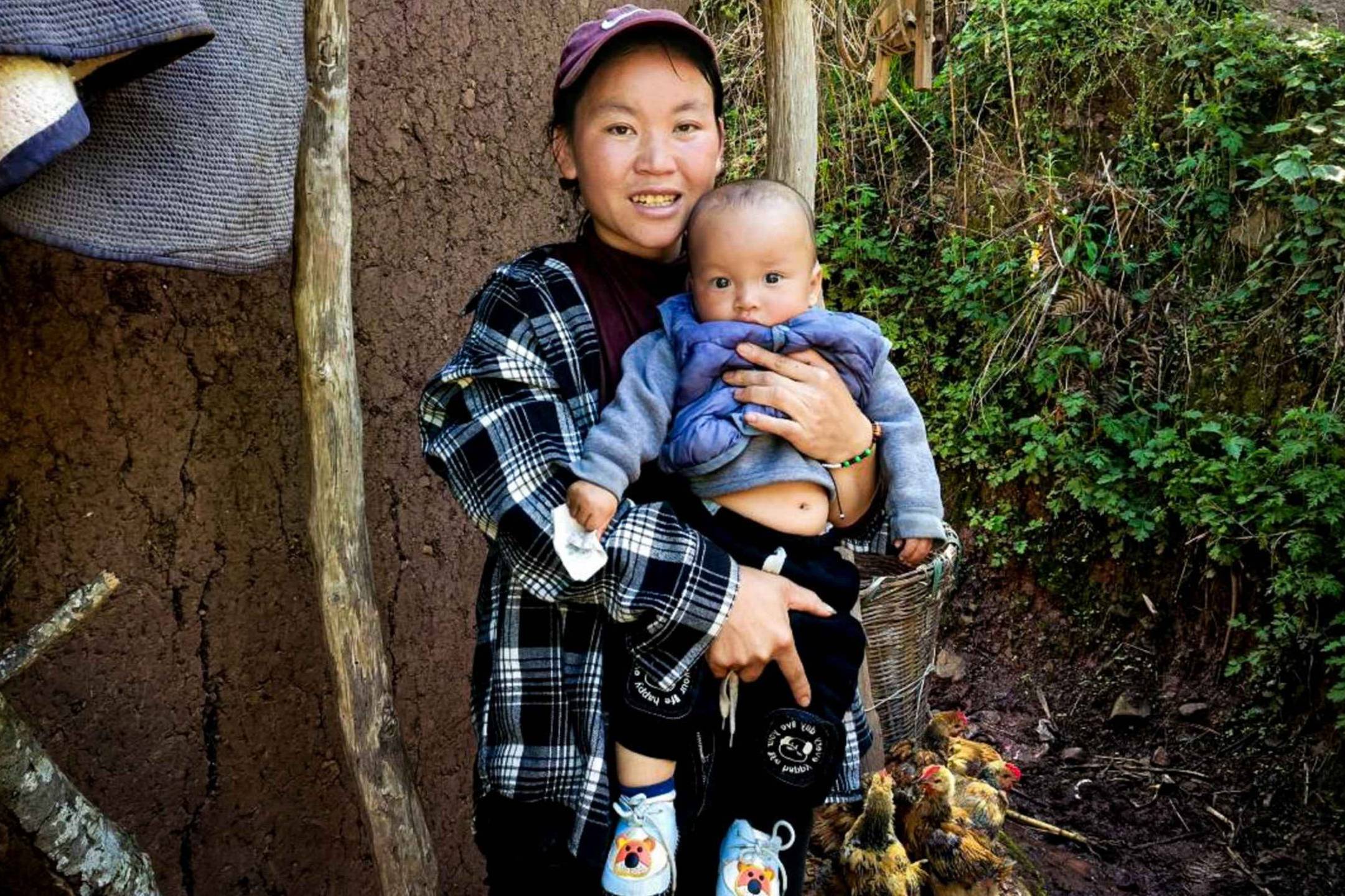 A woman cradles a baby in front of a rustic hut, showcasing a tender moment in a natural setting.