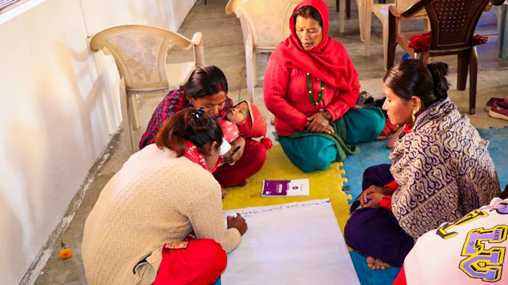 Women gathered on the floor, discussing ideas. One of the women cradles her child while another woman is writing something down on a big piece of paper.