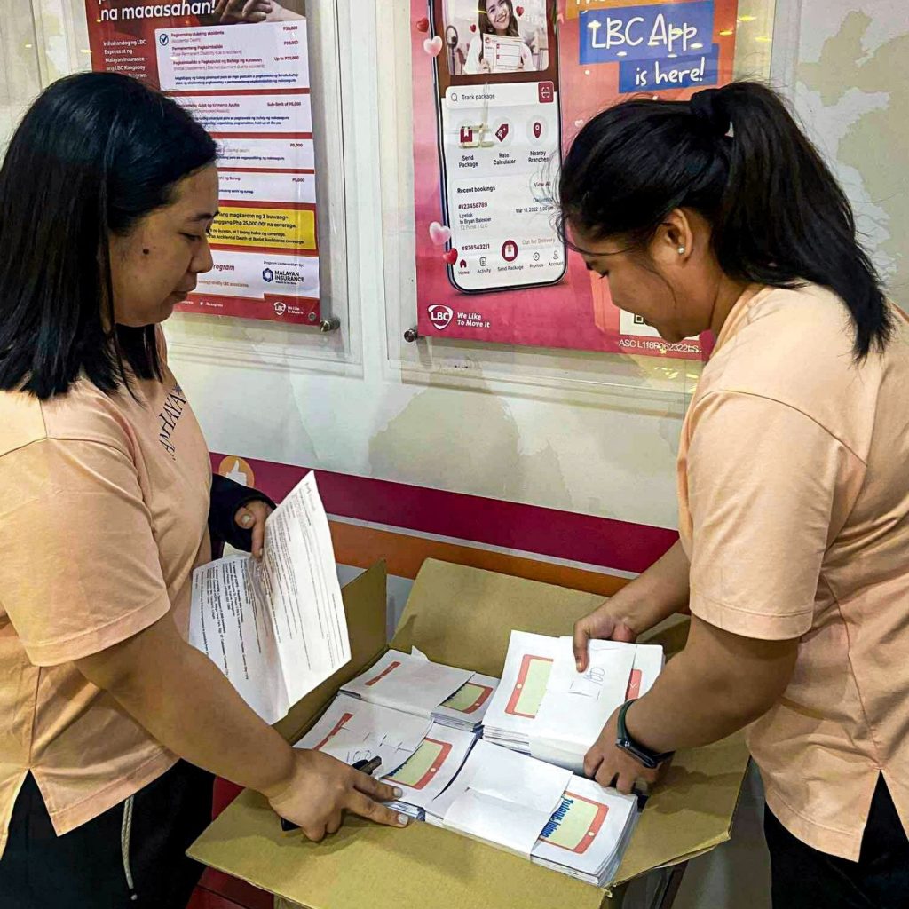 Two women in orange shirts are organizing and sorting through a stack of papers on a table.
