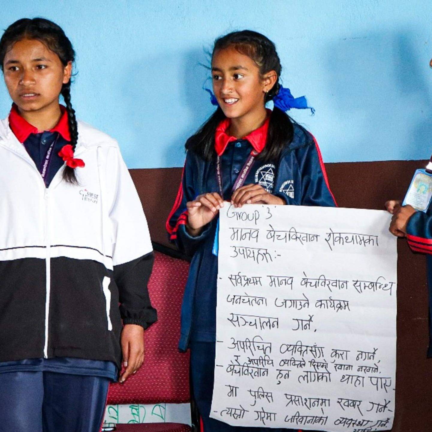 Three girls in school uniforms proudly hold up a sign, smiling and posing together in a bright school environment.