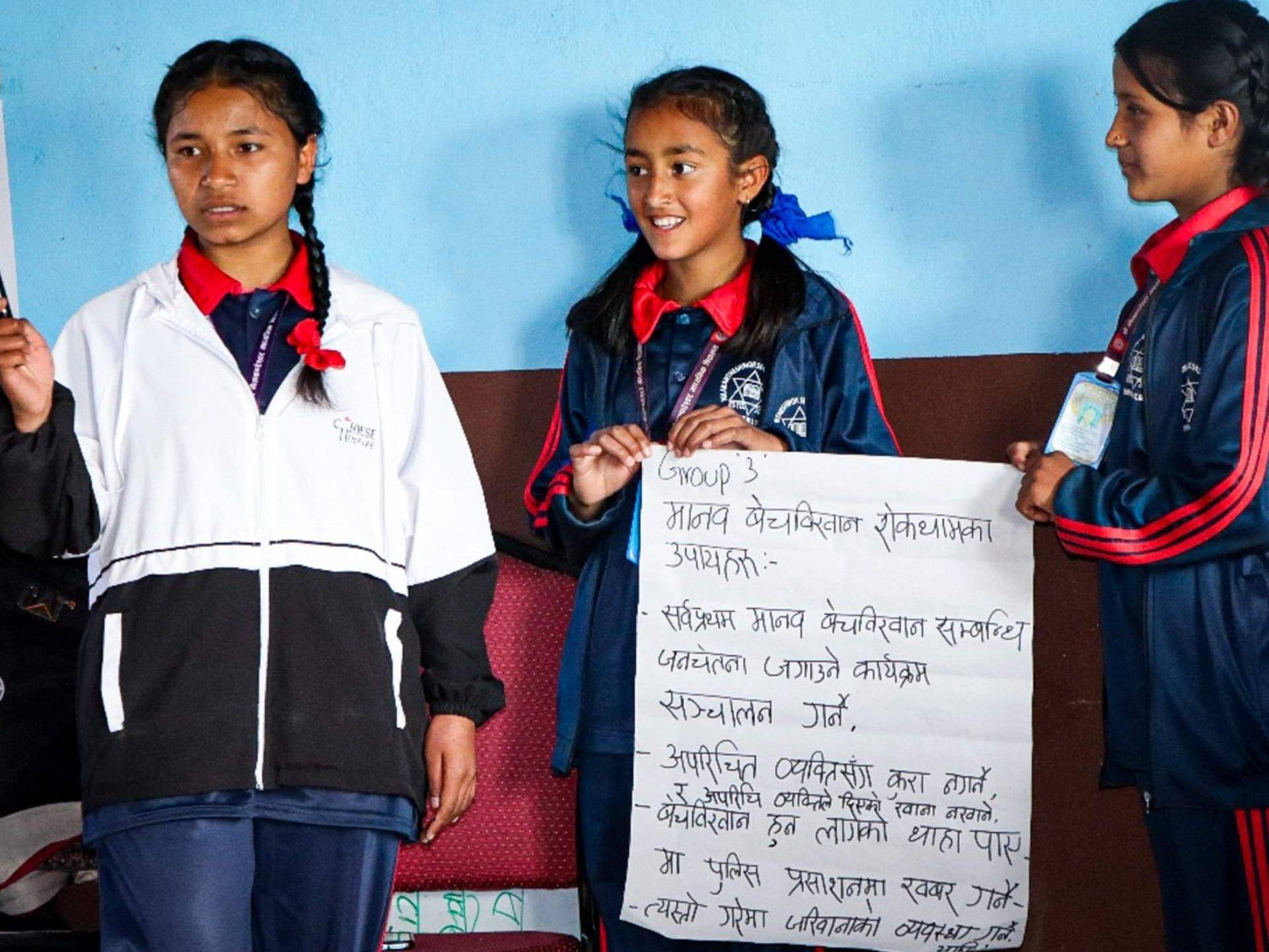 Three girls in school uniforms proudly hold up a sign, smiling and posing together in a bright school environment.