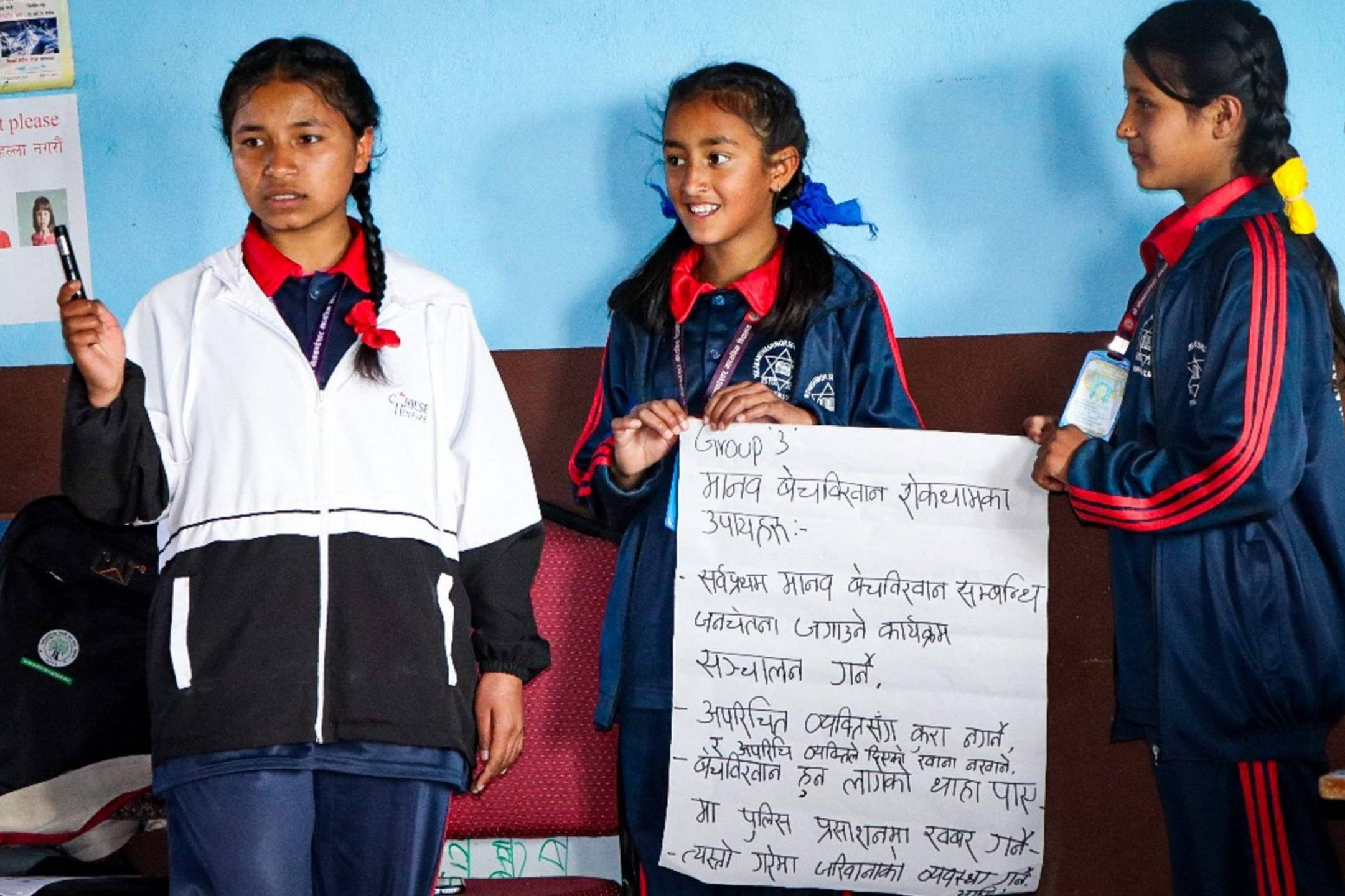 Three girls in school uniforms proudly hold up a sign, smiling and posing together in a bright school environment.