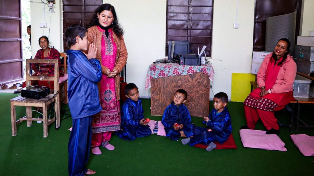 A woman wearing a pink dress stands before a group of children, who are attentively looking at her.