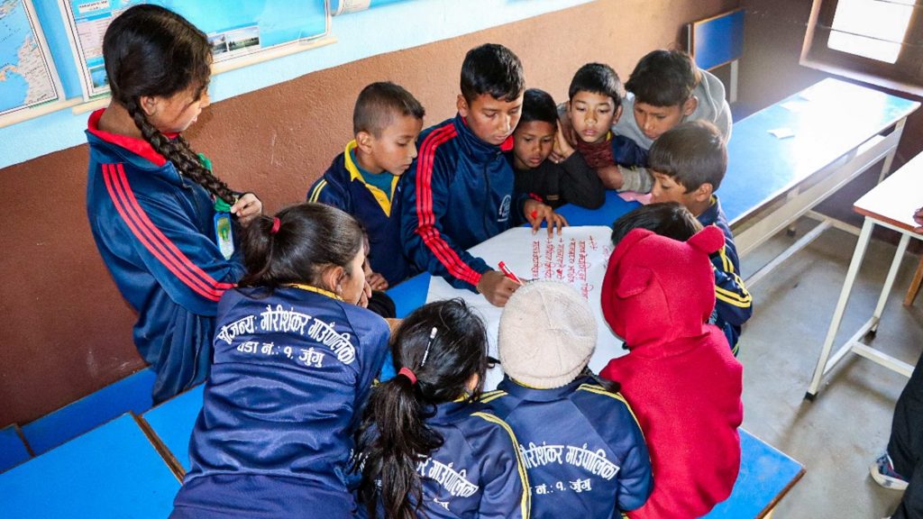 Several children in a classroom focused on a paper, engaged in discussion.