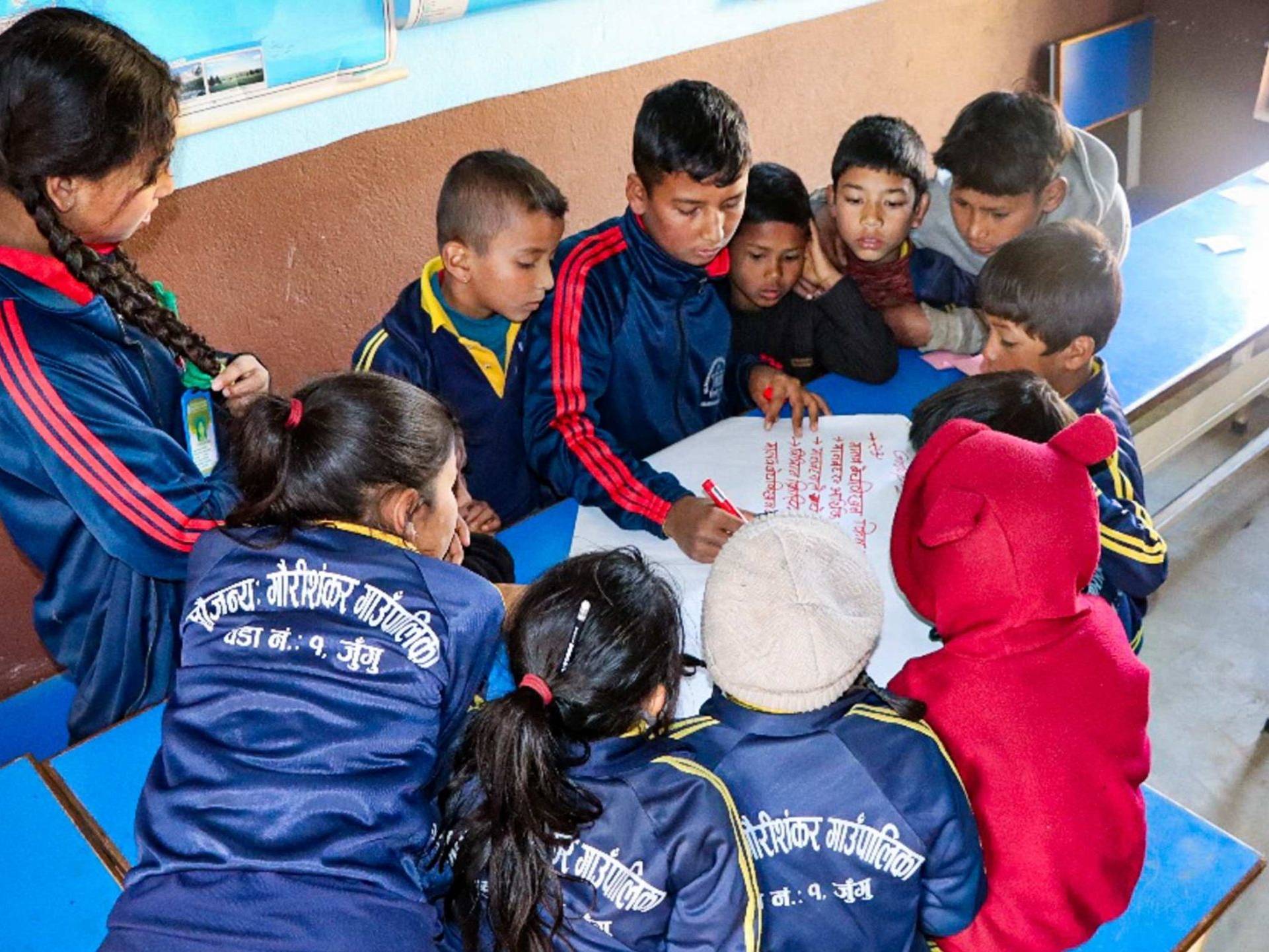 Several children in a classroom focused on a paper, engaged in discussion.