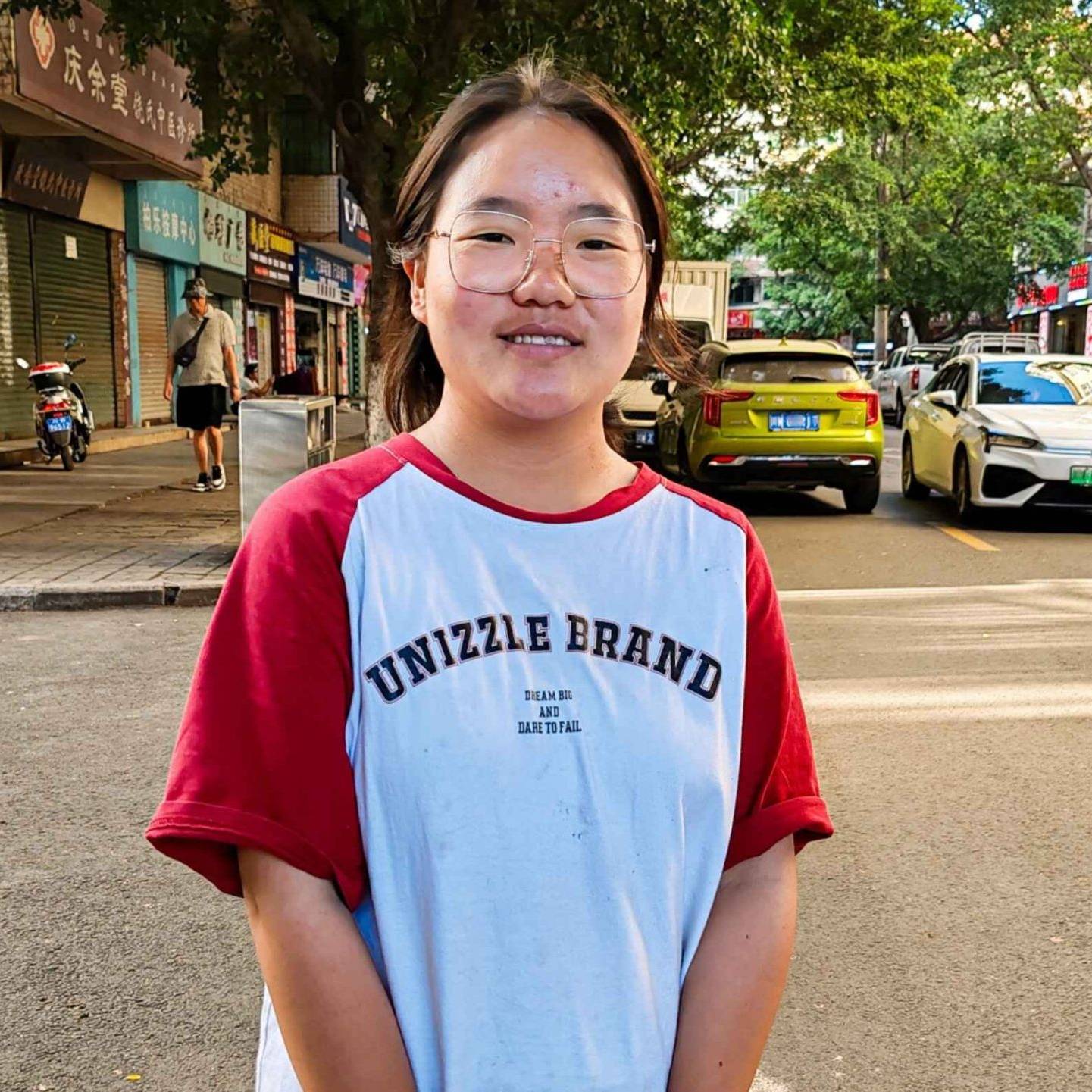 A young woman stands at a street, dressed casually and smiling.