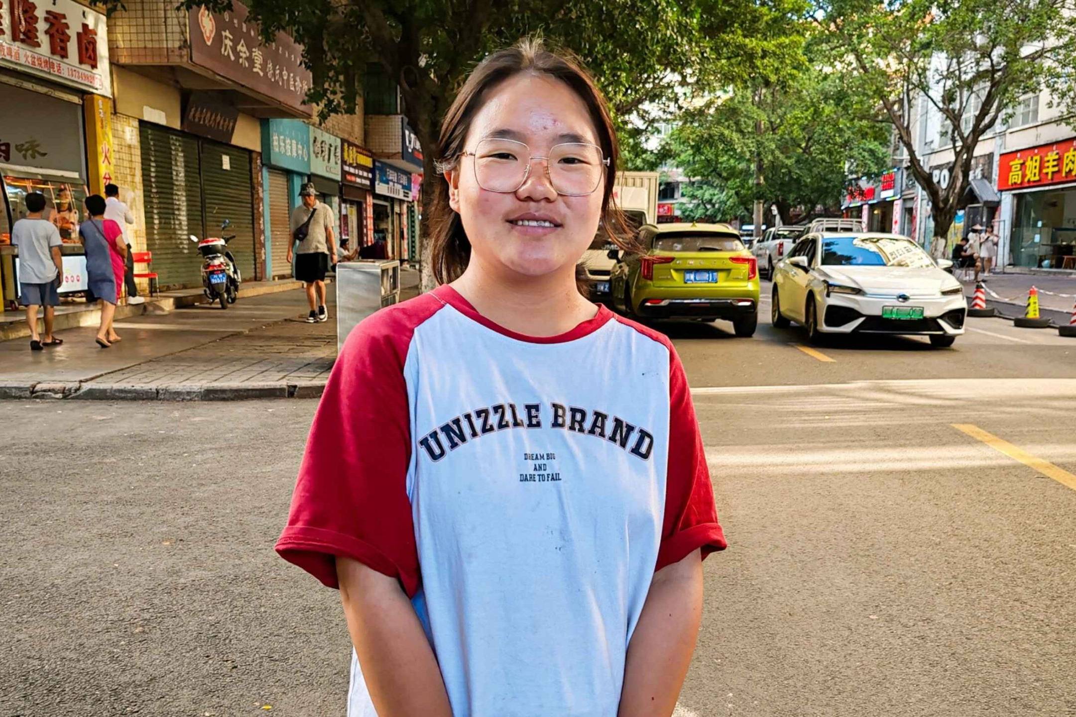 A young woman stands at a street, dressed casually and smiling.