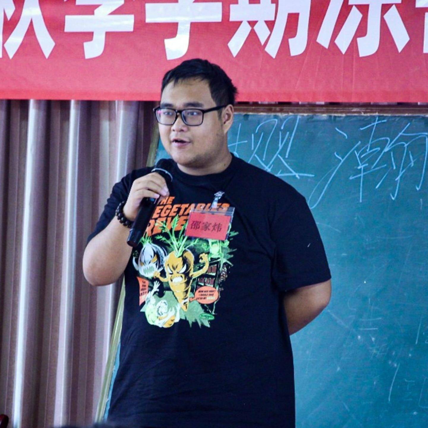 A man in a black shirt speaks into a microphone, engaging with an audience during a presentation or event.