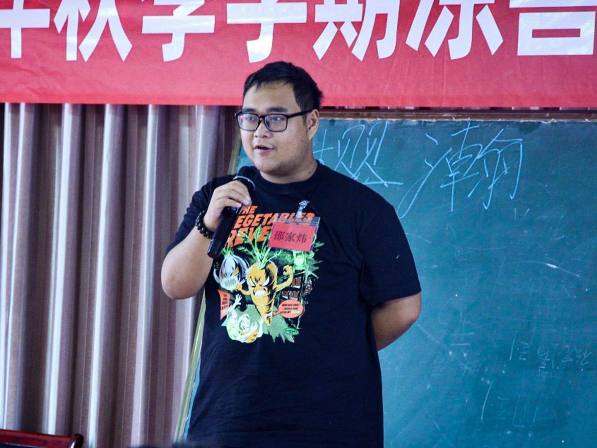 A man in a black shirt speaks into a microphone, engaging with an audience during a presentation or event.