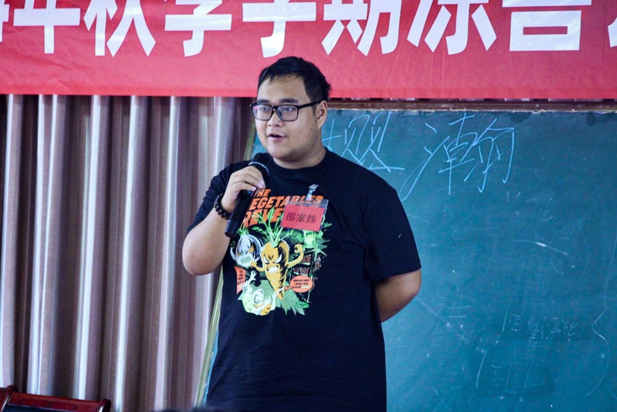 A man in a black shirt speaks into a microphone, engaging with an audience during a presentation or event.