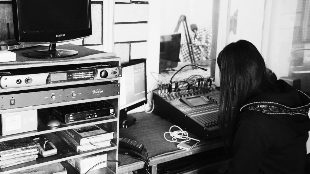 A woman sits at a desk with a computer and a mixer, focused on her work in a well-lit office environment.
