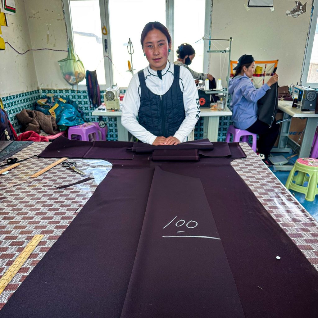 A person stands behind a large table with dark fabric labeled "100." In the background, two people sit sewing. The workspace has a casual, industrious feel.