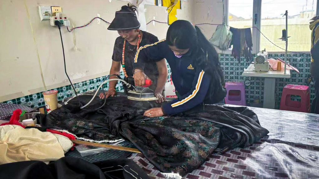 Two women in a workshop are focused on steaming a black fabric on a table. Sewing tools and machines surround them, creating a busy and industrious atmosphere.