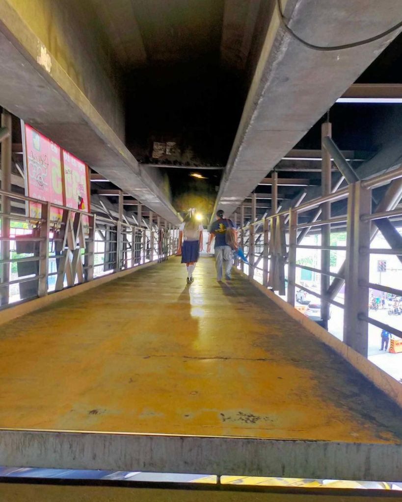 People walk on a vibrant yellow pedestrian bridge under an industrial structure. The scene conveys a sense of urban movement and modernity.