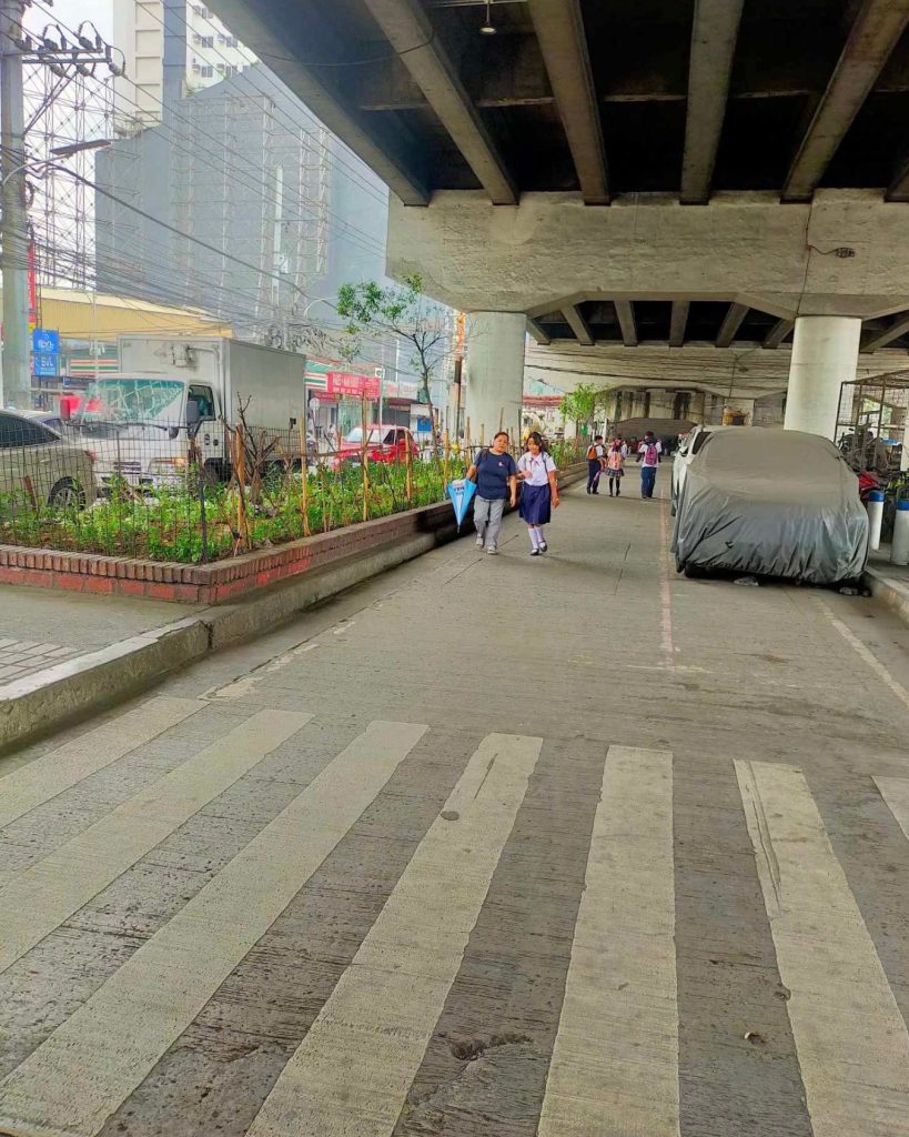 Pedestrian path under a bridge with a crosswalk in foreground. People walk along the path, flanked by greenery on the left and parked cars on the right. Urban setting.