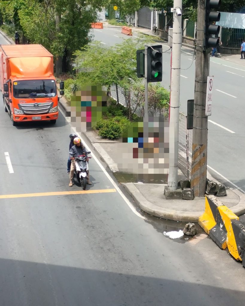 An orange truck and a motorbike with two riders stop at a green light on a city street. Sidewalk has trees, bushes, and blurry figures. Bright, sunny day.