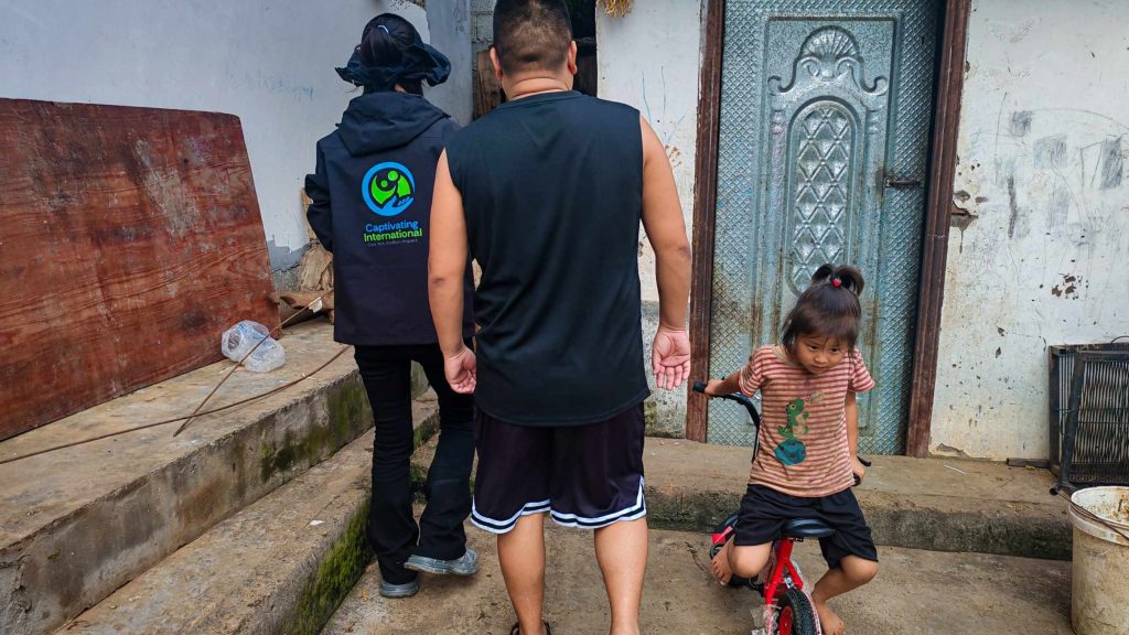 A man and a child with a red bike walk toward a worn door beside a person in a "Captivating International" jacket; a rural setting.