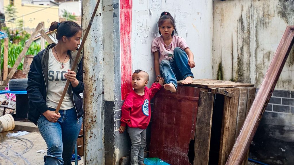 A young woman holds a stick, standing beside two children. One child is seated on a wooden box, the other is standing beside her.