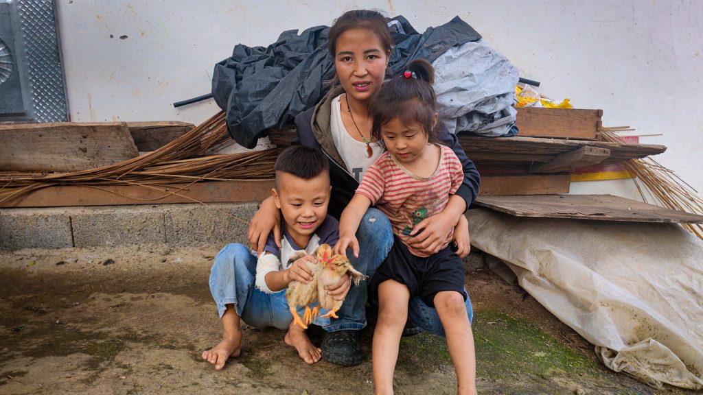 A woman and two children sit on the ground, smiling warmly. The boy holds a chicken, and the girl leans close. They're in front of stacked wooden boards.