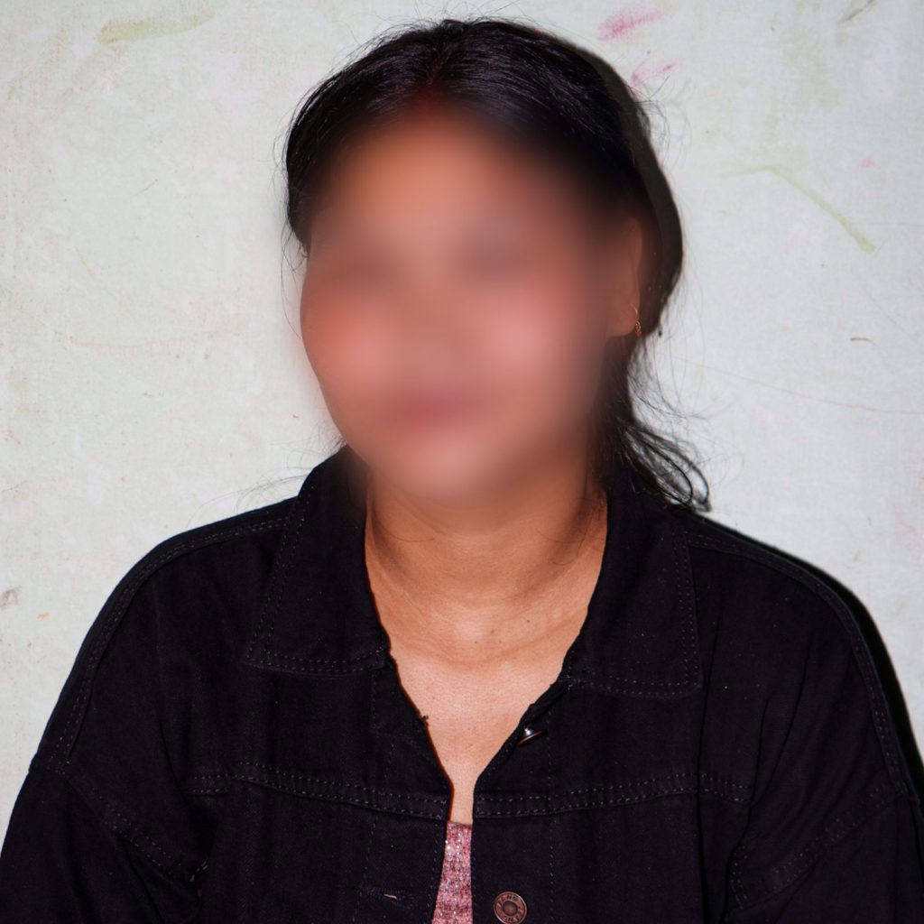 A person with blurred features wearing a black jacket over a pink top stands against a light-colored wall.
