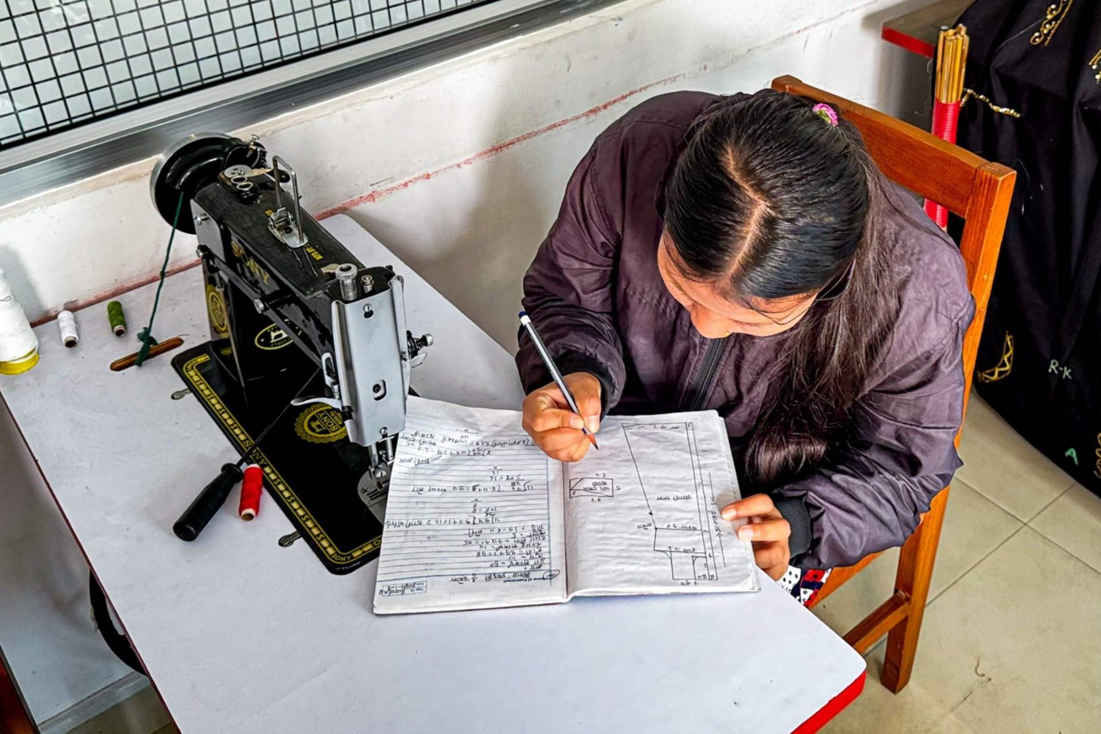 A woman sits at a table with a vintage sewing machine, attentively writing in a notebook.