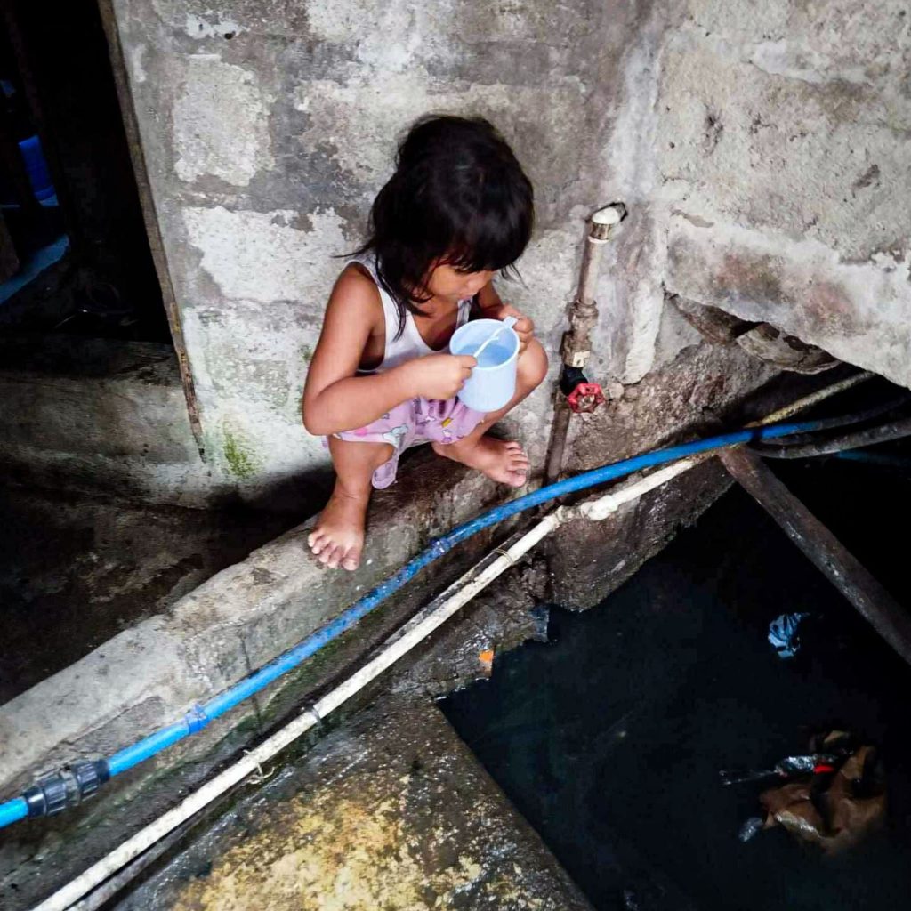 A young girl is perched on a concrete ledge with a blue cup, looking down into a dark, cluttered water hole in a worn, gray industrial setting.