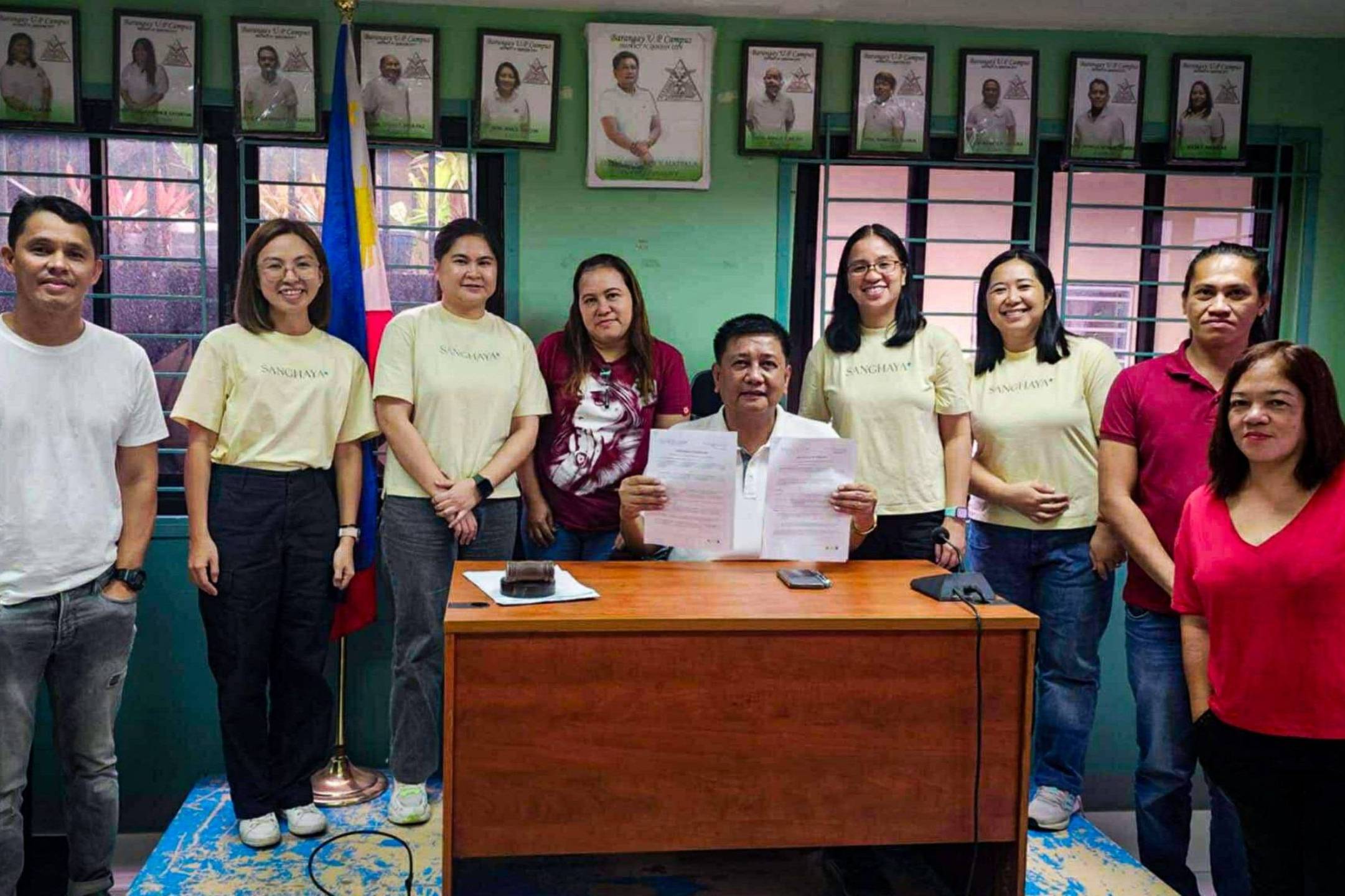 Eight people stand in a room with green walls and framed photos. A man sits at a desk holding up documents, creating a celebratory tone.