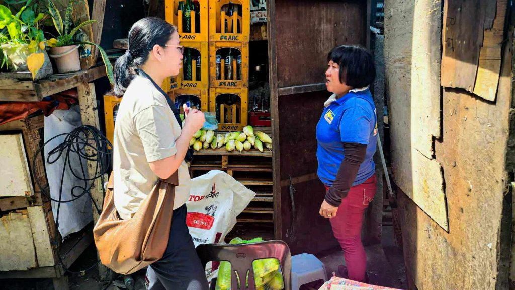 A woman in a beige shirt talks to a market vendor in a blue shirt. Bananas and bottles are displayed around them. 