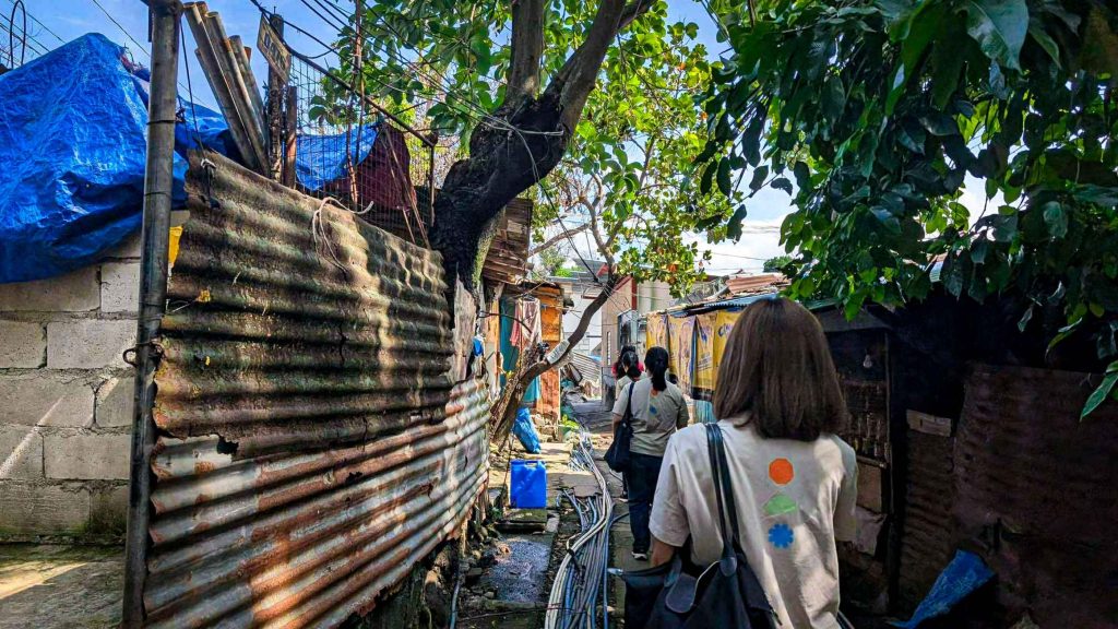 A narrow alleyway lined with corrugated metal sheets and trees, people walk through under dappled light. 