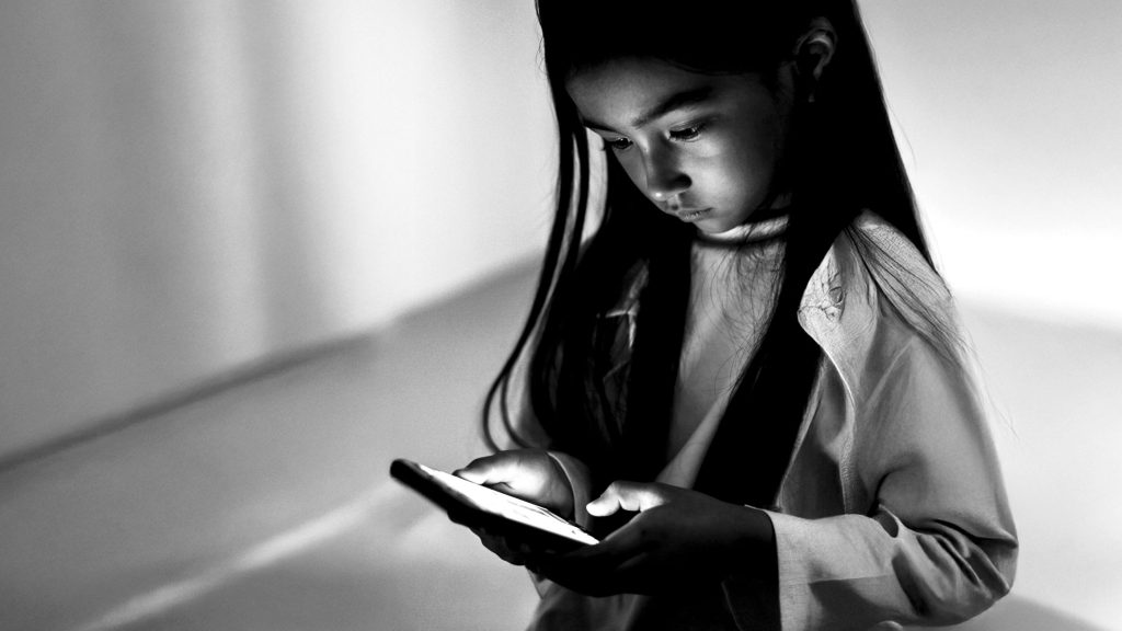 A young girl with long hair intently looks at a glowing smartphone in a dimly lit room. The black-and-white image conveys focus and curiosity.