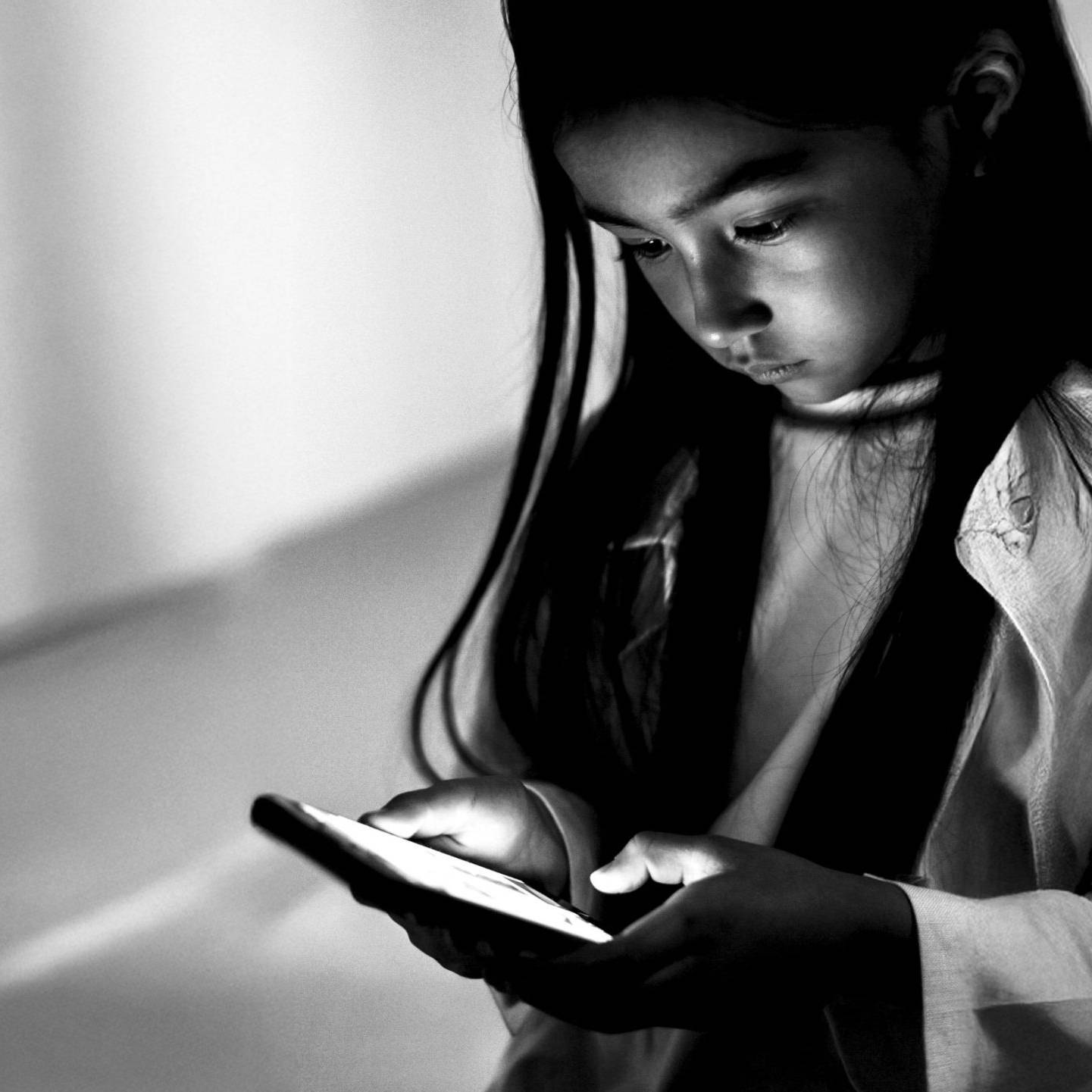 A young girl with long hair intently looks at a glowing smartphone in a dimly lit room. The black-and-white image conveys focus and curiosity.