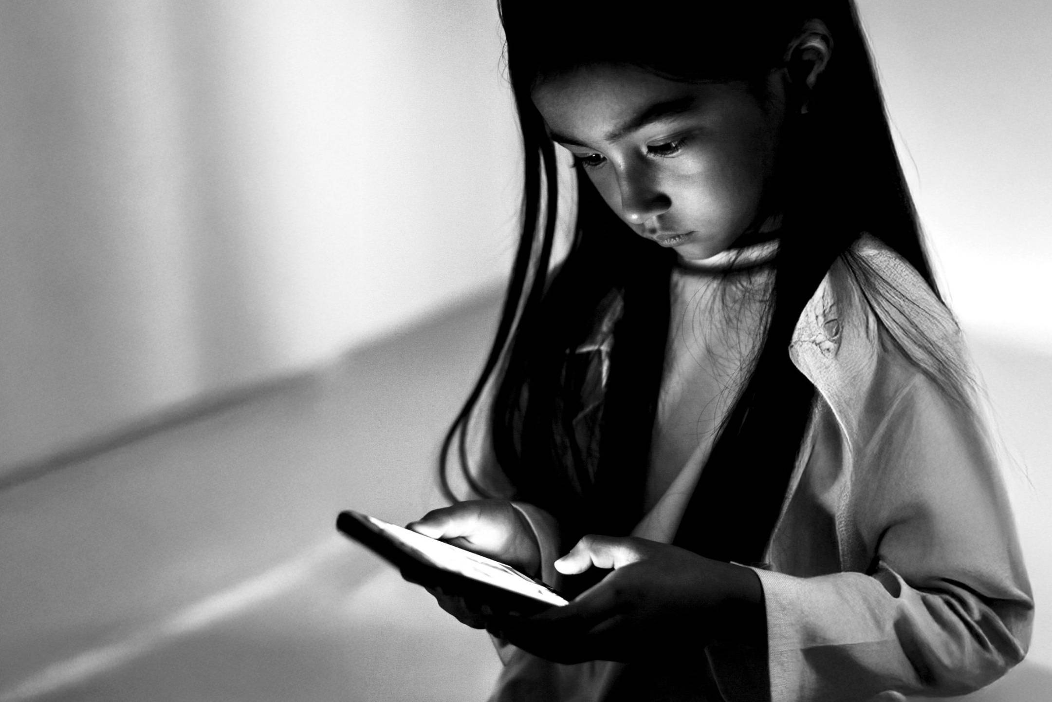 A young girl with long hair intently looks at a glowing smartphone in a dimly lit room. The black-and-white image conveys focus and curiosity.