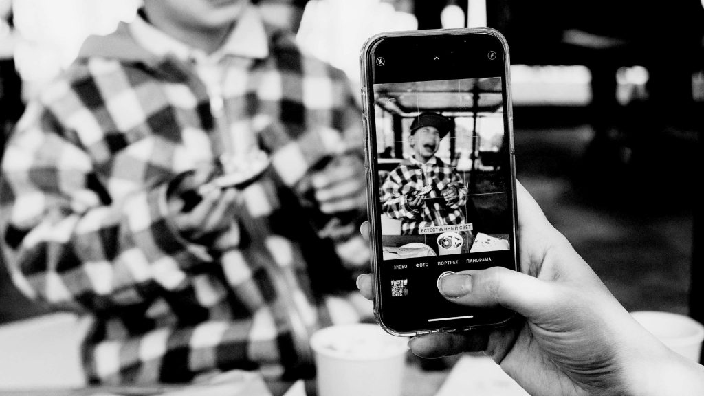 Black and white image of a person taking a photo of a child in a checkered jacket using a smartphone. The scene conveys a cheerful and candid moment.
