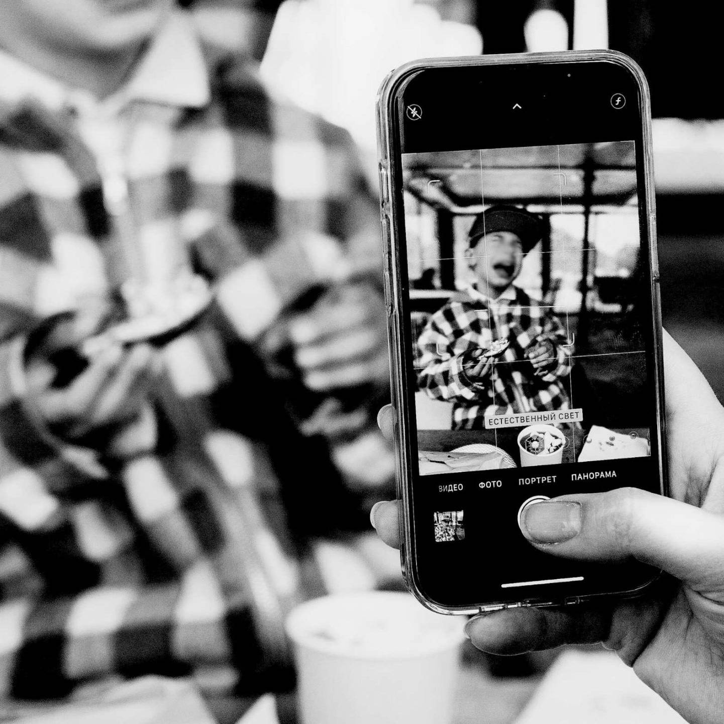 Black and white image of a person taking a photo of a child in a checkered jacket using a smartphone. The scene conveys a cheerful and candid moment.