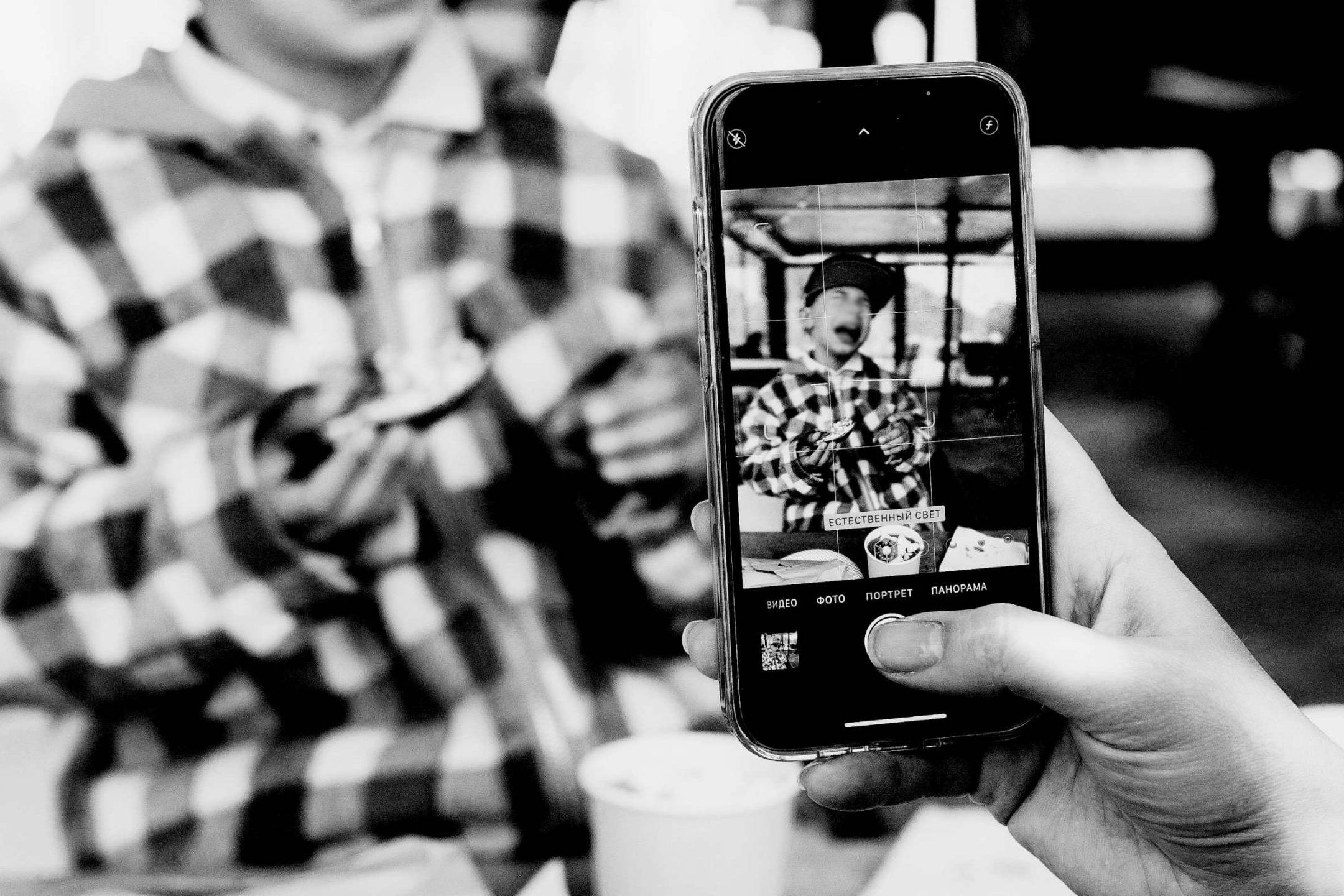 Black and white image of a person taking a photo of a child in a checkered jacket using a smartphone. The scene conveys a cheerful and candid moment.