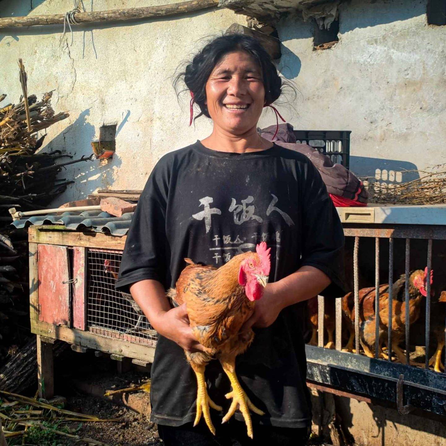 A smiling woman holds a chicken in front of a rustic coop, with chickens inside. A child stands nearby.