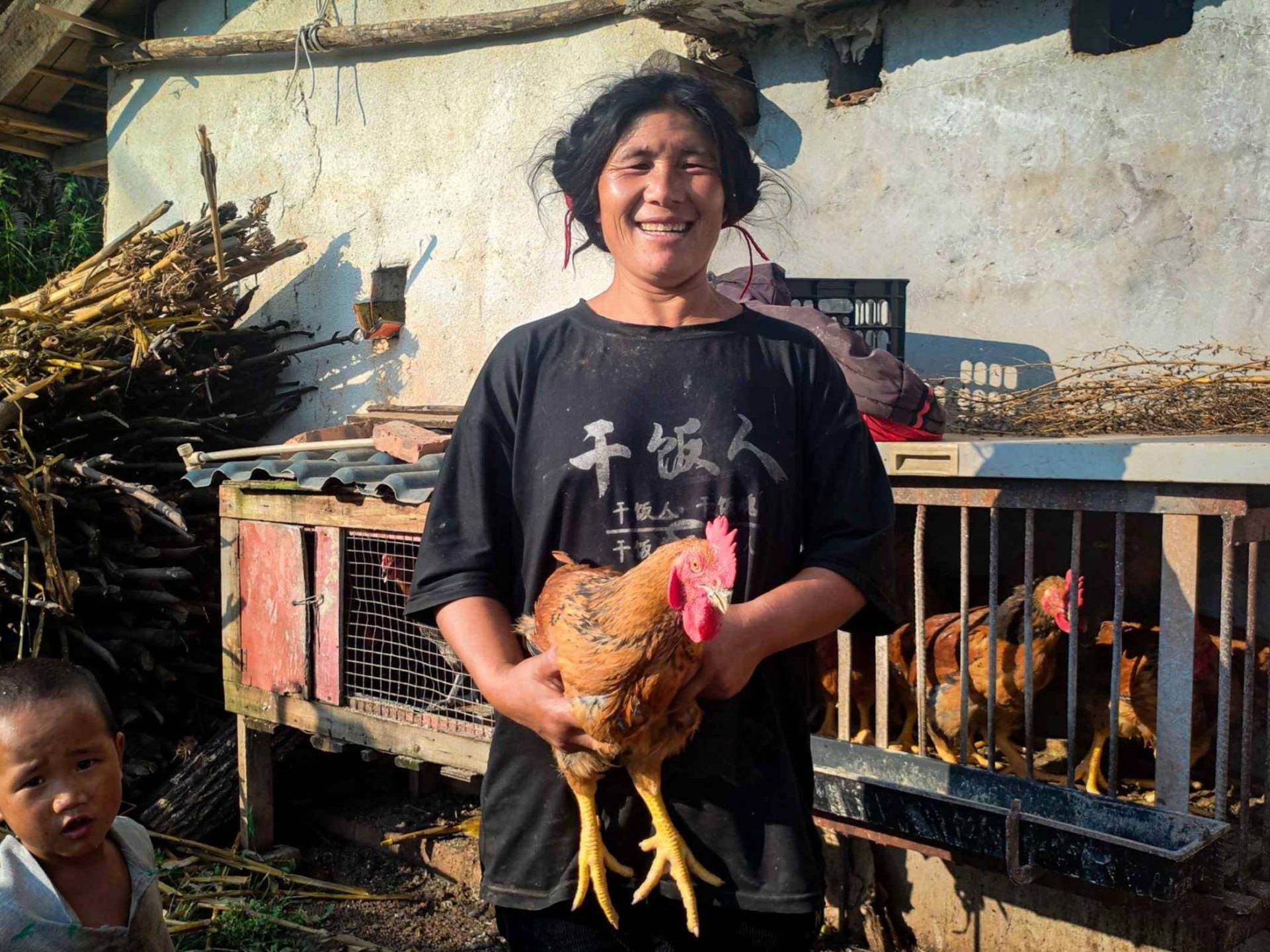 A smiling woman holds a chicken in front of a rustic coop, with chickens inside. A child stands nearby.