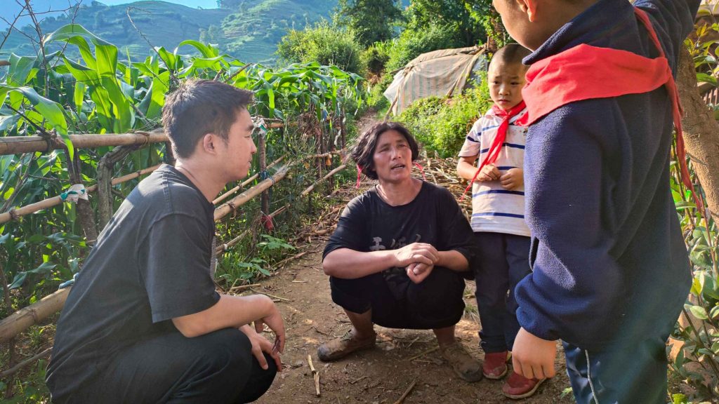 A woman crouches, talking with a man and two children on a dirt path bordered by lush greenery and mountains